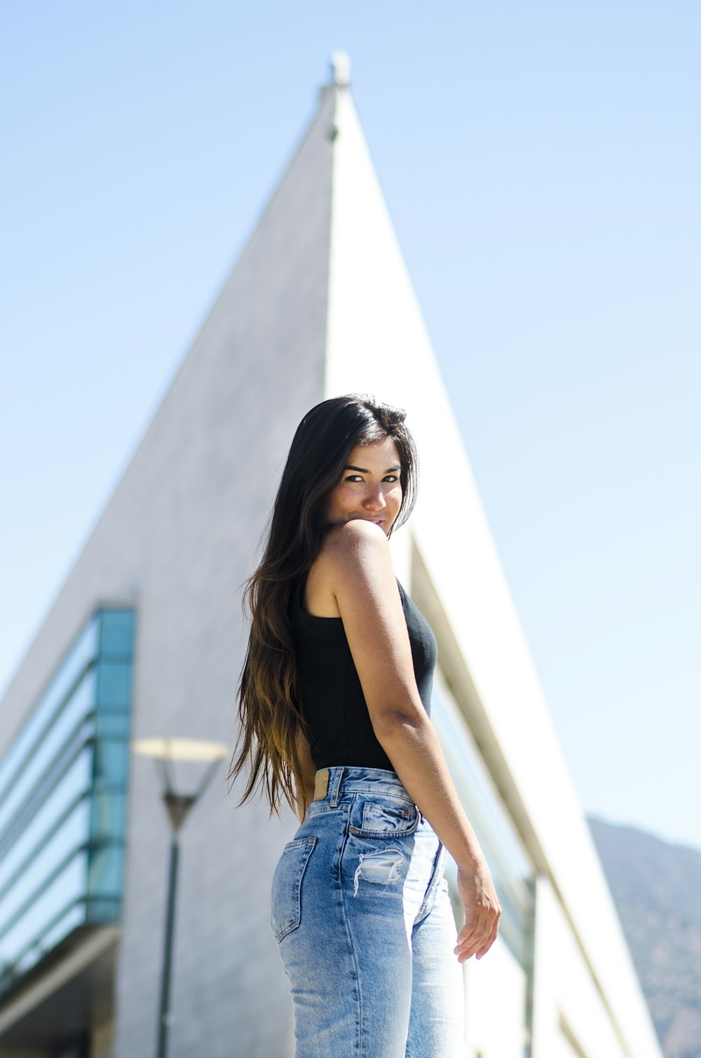 low-angle photo of woman wearing black tank top and blue denim bottoms with view of triangular gray building