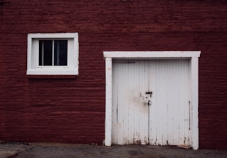 A red brick wall features a small, single-window with white trim on the left side and a large, white, wooden door with a lock in the center. The door has visible signs of weathering and the ground below it is paved with patches of dirt.