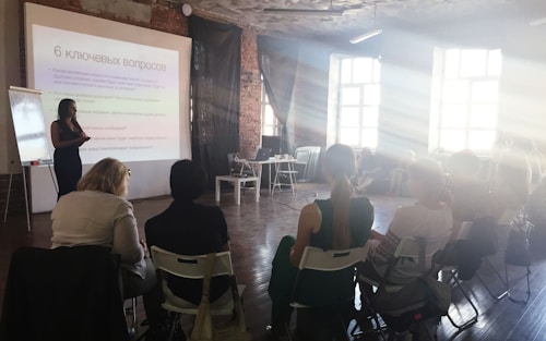 A group of people are seated in a room with exposed brick walls and large windows. Rays of sunlight illuminate the space. A presenter stands in front of a projection screen displaying a slide in Russian, and a flipchart is nearby.