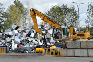 A yellow excavator is positioned in front of a large pile of metallic and electronic waste, including old appliances and scrap metal. Behind the scene, there are trees and a cloudy sky, and concrete barriers are placed in the foreground. The excavator appears to be in the process of sorting or collecting the waste.