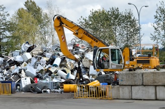 A yellow excavator is positioned in front of a large pile of metallic and electronic waste, including old appliances and scrap metal. Behind the scene, there are trees and a cloudy sky, and concrete barriers are placed in the foreground. The excavator appears to be in the process of sorting or collecting the waste.