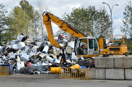 A yellow excavator is positioned in front of a large pile of metallic and electronic waste, including old appliances and scrap metal. Behind the scene, there are trees and a cloudy sky, and concrete barriers are placed in the foreground. The excavator appears to be in the process of sorting or collecting the waste.