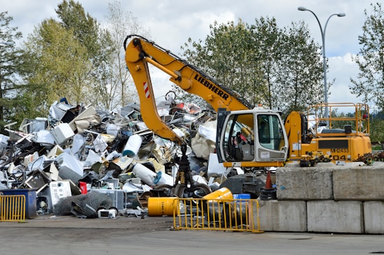 A yellow excavator is positioned in front of a large pile of metallic and electronic waste, including old appliances and scrap metal. Behind the scene, there are trees and a cloudy sky, and concrete barriers are placed in the foreground. The excavator appears to be in the process of sorting or collecting the waste.