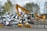 A yellow excavator is positioned in front of a large pile of metallic and electronic waste, including old appliances and scrap metal. Behind the scene, there are trees and a cloudy sky, and concrete barriers are placed in the foreground. The excavator appears to be in the process of sorting or collecting the waste.