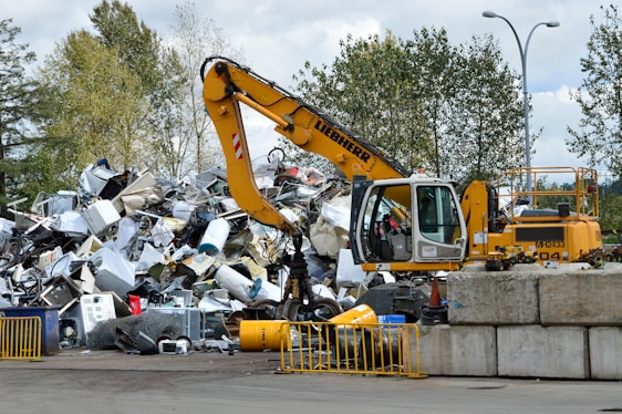 A yellow excavator is positioned in front of a large pile of metallic and electronic waste, including old appliances and scrap metal. Behind the scene, there are trees and a cloudy sky, and concrete barriers are placed in the foreground. The excavator appears to be in the process of sorting or collecting the waste.