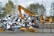 A yellow excavator is positioned in front of a large pile of metallic and electronic waste, including old appliances and scrap metal. Behind the scene, there are trees and a cloudy sky, and concrete barriers are placed in the foreground. The excavator appears to be in the process of sorting or collecting the waste.
