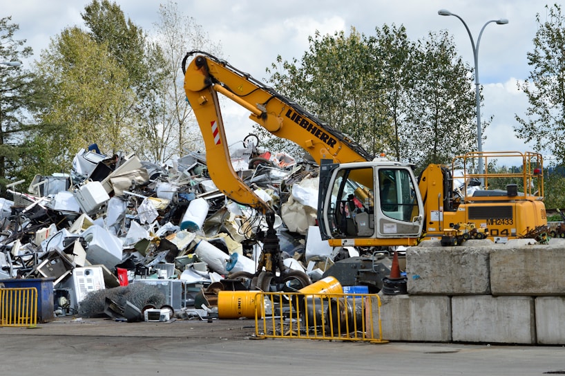 A yellow excavator is positioned in front of a large pile of metallic and electronic waste, including old appliances and scrap metal. Behind the scene, there are trees and a cloudy sky, and concrete barriers are placed in the foreground. The excavator appears to be in the process of sorting or collecting the waste.