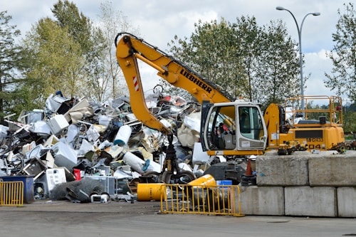 A yellow excavator is positioned in front of a large pile of metallic and electronic waste, including old appliances and scrap metal. Behind the scene, there are trees and a cloudy sky, and concrete barriers are placed in the foreground. The excavator appears to be in the process of sorting or collecting the waste.