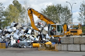 A yellow excavator is positioned in front of a large pile of metallic and electronic waste, including old appliances and scrap metal. Behind the scene, there are trees and a cloudy sky, and concrete barriers are placed in the foreground. The excavator appears to be in the process of sorting or collecting the waste.