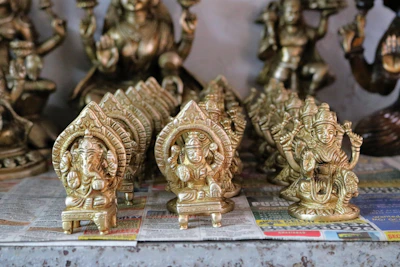 Several small intricately designed brass statues are placed on a surface covered with newspaper. The statues are mostly of Hindu deities, showcasing details such as ornate crowns and intricate carvings. The statues are arranged in rows, with larger statues in the background.