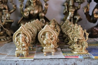 Several small intricately designed brass statues are placed on a surface covered with newspaper. The statues are mostly of Hindu deities, showcasing details such as ornate crowns and intricate carvings. The statues are arranged in rows, with larger statues in the background.