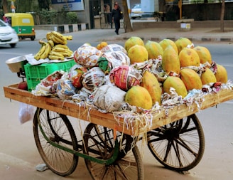 A wooden cart filled with ripe papayas and bananas is parked on the side of a busy road. The papayas are wrapped in newspaper, and there are some exposed bananas on a green plastic crate. Vehicles are seen in the background, including a green auto-rickshaw and a white car, indicating a bustling street.