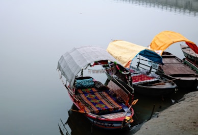 Three ornately decorated boats are docked along a calm waterway. Each boat features a colorful canopy; the canopies are white, yellow, and orange, offering shelter to the passengers. The foreground boat displays a vibrant, patterned seating area with detailed ornamentation.