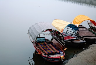 Three ornately decorated boats are docked along a calm waterway. Each boat features a colorful canopy; the canopies are white, yellow, and orange, offering shelter to the passengers. The foreground boat displays a vibrant, patterned seating area with detailed ornamentation.