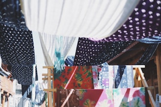 Colorful fabrics with various patterns, such as polka dots and floral designs, are hanging on wooden racks and lines. They create a vibrant tapestry of colors and textures, with the sunlight casting dynamic shadows between them.