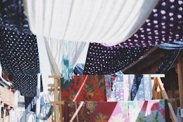 Colorful fabrics with various patterns, such as polka dots and floral designs, are hanging on wooden racks and lines. They create a vibrant tapestry of colors and textures, with the sunlight casting dynamic shadows between them.