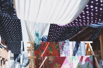 Colorful fabrics with various patterns, such as polka dots and floral designs, are hanging on wooden racks and lines. They create a vibrant tapestry of colors and textures, with the sunlight casting dynamic shadows between them.