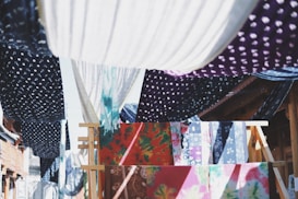 Colorful fabrics with various patterns, such as polka dots and floral designs, are hanging on wooden racks and lines. They create a vibrant tapestry of colors and textures, with the sunlight casting dynamic shadows between them.