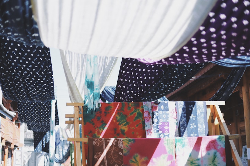 Colorful fabrics with various patterns, such as polka dots and floral designs, are hanging on wooden racks and lines. They create a vibrant tapestry of colors and textures, with the sunlight casting dynamic shadows between them.