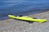 A bright green kayak is lying on a rocky beach, with clear blue water gently lapping the shore. The kayak has black paddles placed beside it, and there is minimal vegetation or debris around.