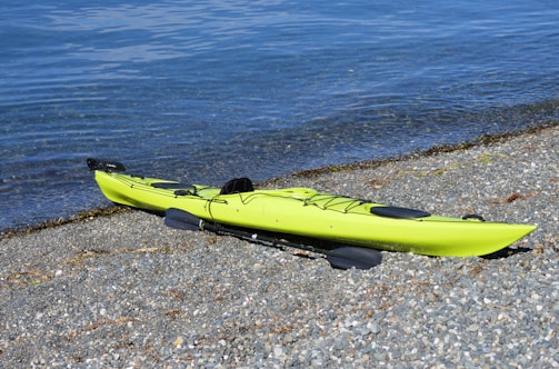 A bright green kayak is lying on a rocky beach, with clear blue water gently lapping the shore. The kayak has black paddles placed beside it, and there is minimal vegetation or debris around.