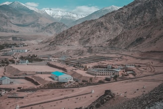 A high-altitude mountainous region with snow-capped peaks in the background. Near the base of the mountain, several modern buildings and structures are dispersed throughout the arid and rocky landscape. The buildings have varied architecture, with one noticeable structure having a distinct blue roof. There is minimal vegetation, with a few patches of greenery near the buildings.