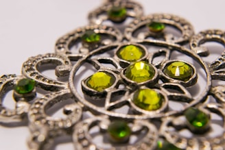 A close-up image of an intricately designed metal ornament featuring several green gemstones. The pattern consists of circular and curved metalwork surrounding the gems, creating a detailed and ornate appearance.