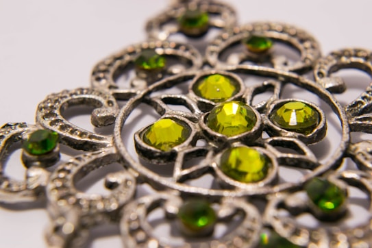 A close-up image of an intricately designed metal ornament featuring several green gemstones. The pattern consists of circular and curved metalwork surrounding the gems, creating a detailed and ornate appearance.