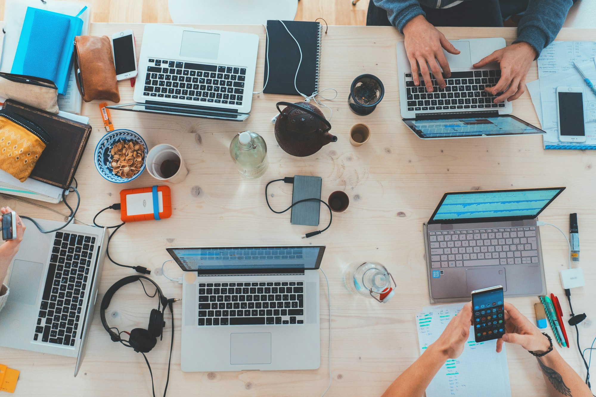 Ein Holztisch voller Laptops, Snacks, Handys und Kopfhörer, an dem mehrere Leute arbeiten.