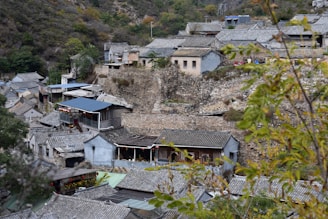 A village nestled in a mountainous area features traditional stone and tile-roofed houses. The buildings are closely arranged on a rocky slope, with some greenery and foliage present in the foreground.