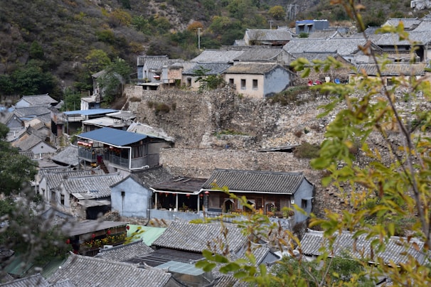A village nestled in a mountainous area features traditional stone and tile-roofed houses. The buildings are closely arranged on a rocky slope, with some greenery and foliage present in the foreground.