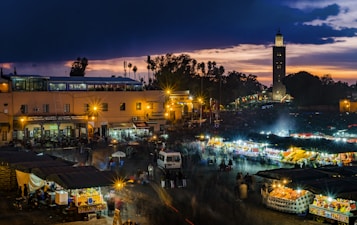 A bustling market scene at dusk, with vibrant stalls illuminated by bright lights against a backdrop of a deep blue and orange sunset sky. The image includes a tall tower in the background, surrounded by trees and silhouetted by the fading light. People are walking around the market, creating a lively and energetic atmosphere.