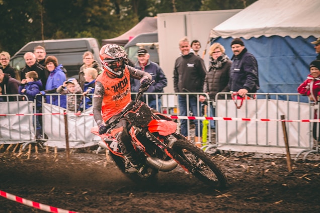 A motocross rider in an orange outfit navigates a dirt track surrounded by spectators. The crowd stands behind metal barriers, and the track is marked with red and white tape. The ground is muddy, and the rider's bike is partially covered in dirt.