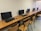 A row of computer workstations with flat-screen monitors placed on wooden desks. Each station includes a keyboard and a mouse, with wooden chairs in front of them. The setting is a neat and orderly computer lab or office space with a plain wall in the background.