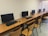 A row of computer workstations with flat-screen monitors placed on wooden desks. Each station includes a keyboard and a mouse, with wooden chairs in front of them. The setting is a neat and orderly computer lab or office space with a plain wall in the background.