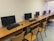 A row of computer workstations with flat-screen monitors placed on wooden desks. Each station includes a keyboard and a mouse, with wooden chairs in front of them. The setting is a neat and orderly computer lab or office space with a plain wall in the background.