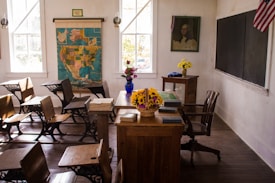 A vintage classroom setting features wooden desks and chairs arranged neatly in rows. A large map of North America hangs on the wall between two windows, and a blackboard is mounted to the right. On the desk at the front of the room, a basket of yellow flowers adds a touch of color. A blue vase with flowers sits on a side table beneath a historical portrait. An American flag is displayed to the right side.