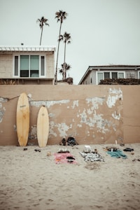 Surfboards lean against a worn, sandy wall near a beach house with large windows and a few tall palm trees in the background. Beach towels with scattered personal items rest on the sand, suggesting a laid-back beach environment.