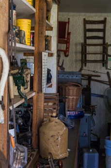 A cluttered interior space with a variety of old items on shelves and scattered around. The shelves contain vintage cans, boxes, and jars. A burlap-covered jug is prominently placed on a counter. Wooden chairs and other furniture pieces are hanging on the back wall, while a wicker basket and metal boxes are visible. The scene conveys a sense of nostalgia and history.