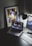 A modern workspace features a laptop displaying a music streaming platform, accompanied by a desk lamp, a keyboard, and a smartphone. A framed abstract art piece with geometric shapes and bold colors is propped against the wall.