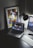 A modern workspace features a laptop displaying a music streaming platform, accompanied by a desk lamp, a keyboard, and a smartphone. A framed abstract art piece with geometric shapes and bold colors is propped against the wall.