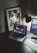 A modern workspace features a laptop displaying a music streaming platform, accompanied by a desk lamp, a keyboard, and a smartphone. A framed abstract art piece with geometric shapes and bold colors is propped against the wall.
