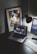 A modern workspace features a laptop displaying a music streaming platform, accompanied by a desk lamp, a keyboard, and a smartphone. A framed abstract art piece with geometric shapes and bold colors is propped against the wall.