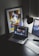 A modern workspace features a laptop displaying a music streaming platform, accompanied by a desk lamp, a keyboard, and a smartphone. A framed abstract art piece with geometric shapes and bold colors is propped against the wall.