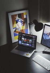 A modern workspace features a laptop displaying a music streaming platform, accompanied by a desk lamp, a keyboard, and a smartphone. A framed abstract art piece with geometric shapes and bold colors is propped against the wall.