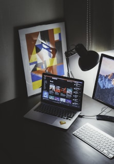 A modern workspace features a laptop displaying a music streaming platform, accompanied by a desk lamp, a keyboard, and a smartphone. A framed abstract art piece with geometric shapes and bold colors is propped against the wall.
