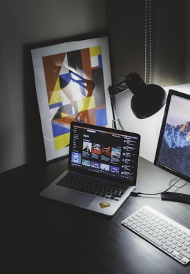 A modern workspace features a laptop displaying a music streaming platform, accompanied by a desk lamp, a keyboard, and a smartphone. A framed abstract art piece with geometric shapes and bold colors is propped against the wall.