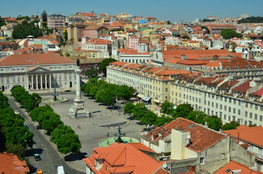 A vibrant cityscape featuring a plaza with a central monument surrounded by historic buildings with red-tiled roofs. The architecture includes classical columns and facades, set amidst lush green trees. The streets are bustling with people and vehicles, showcasing a lively urban atmosphere.