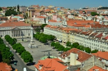 A vibrant cityscape featuring a plaza with a central monument surrounded by historic buildings with red-tiled roofs. The architecture includes classical columns and facades, set amidst lush green trees. The streets are bustling with people and vehicles, showcasing a lively urban atmosphere.