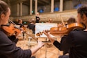 Musicians are seated in a grand concert hall, playing string instruments like violins. The conductor is visible in the background, leading the orchestra. The room is elegant, with high ceilings and ornate architectural details.